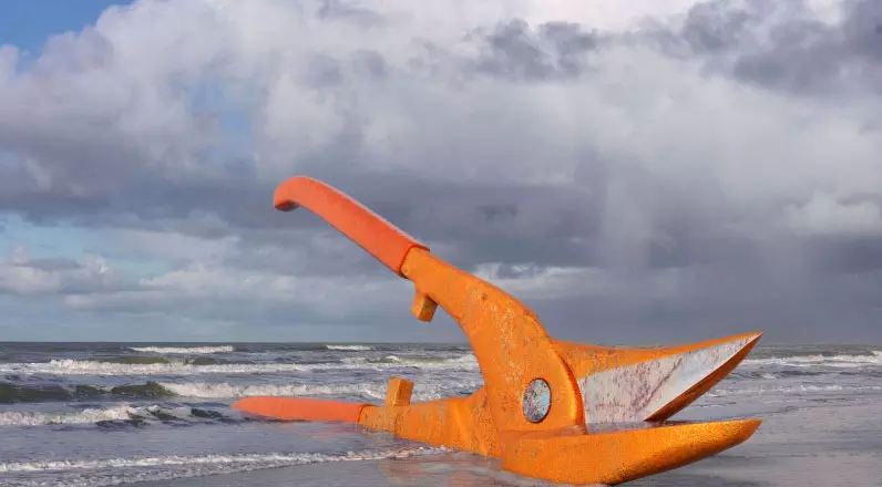 alt="Large orange  industrial shears lying on a beach with waves and a cloudy sky in the background."