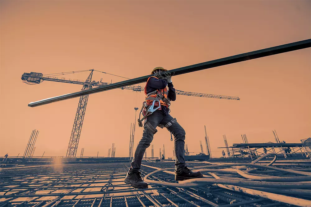 Construction worker carrying a metal beam across a building site with cranes in the background, symbolizing strength and progress.