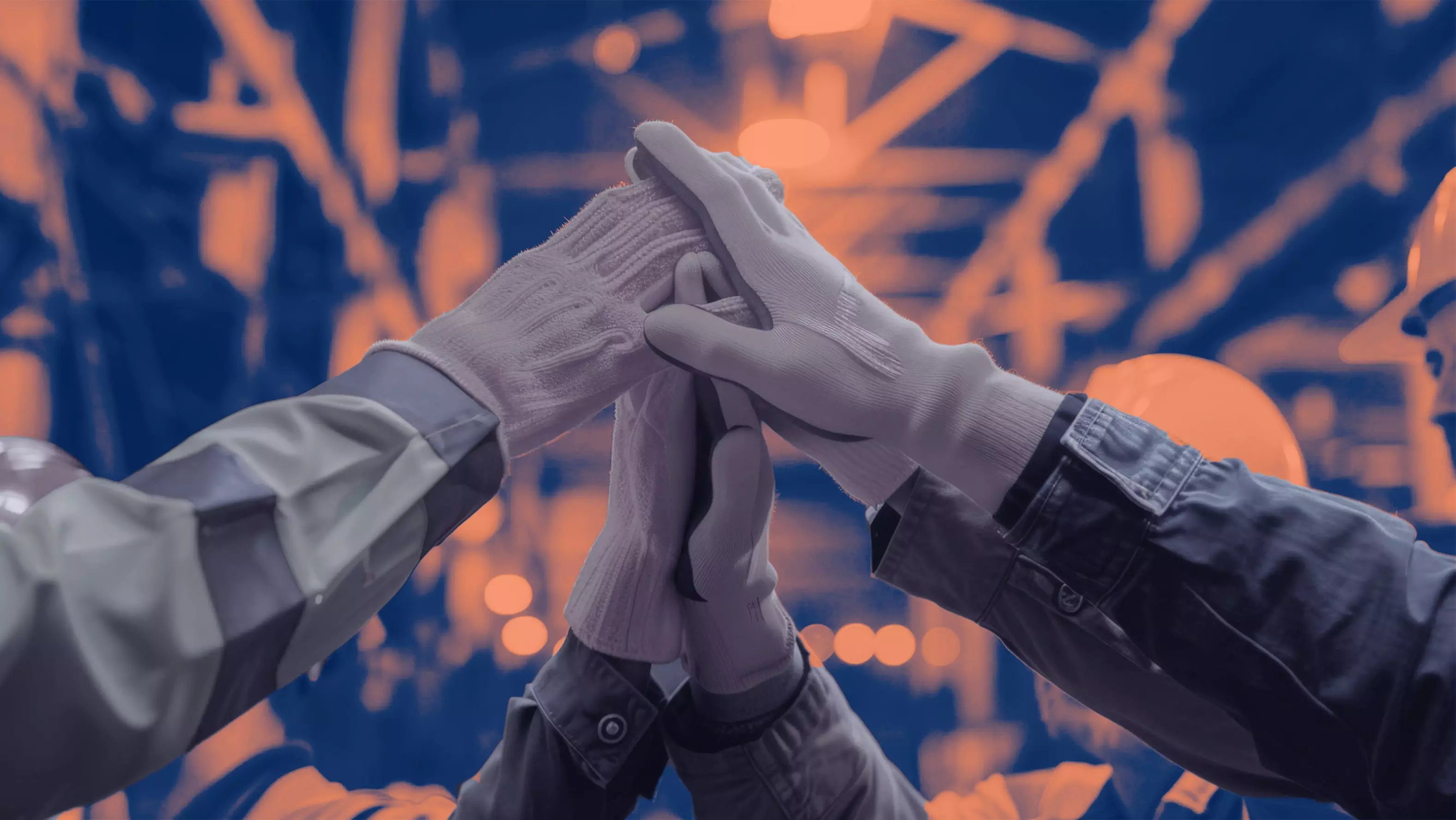 Close-up of construction workers wearing gloves, joining hands together in a teamwork gesture against a blurred industrial background.