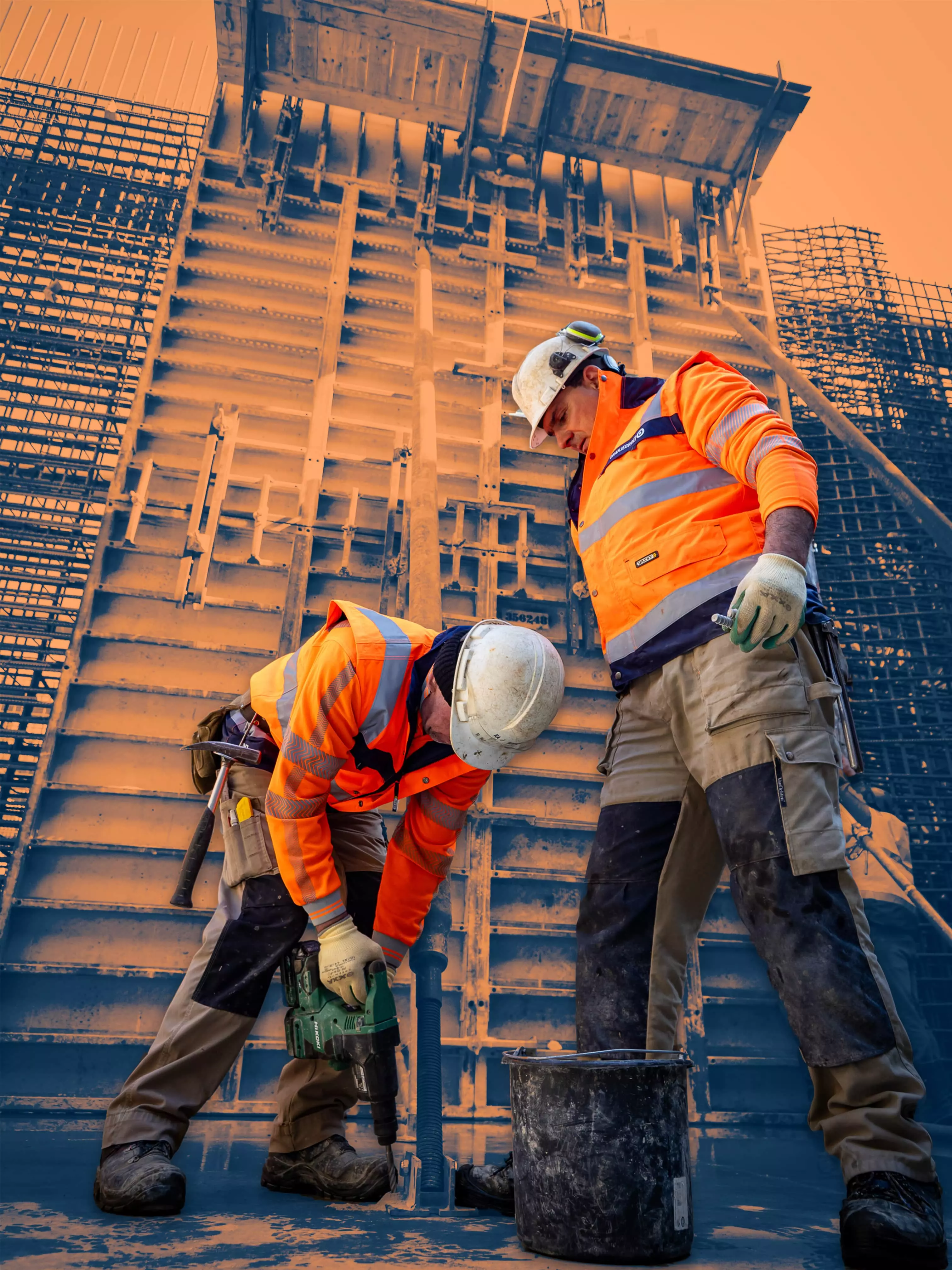 Two construction workers in orange safety vests working near a large vertical formwork structure on a building site.