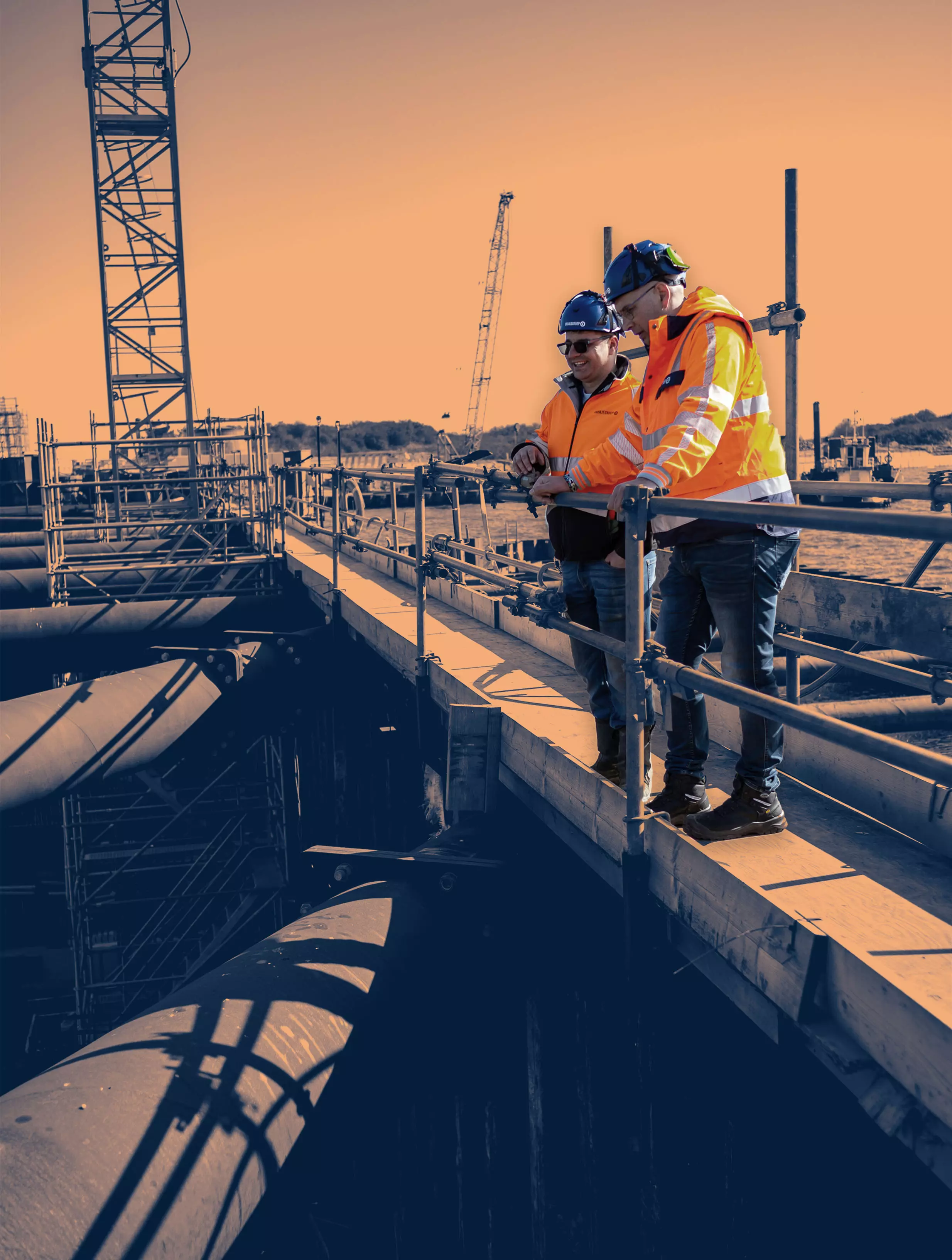 Two construction supervisors in safety gear discussing project progress on-site, standing on a platform above large industrial pipes.

