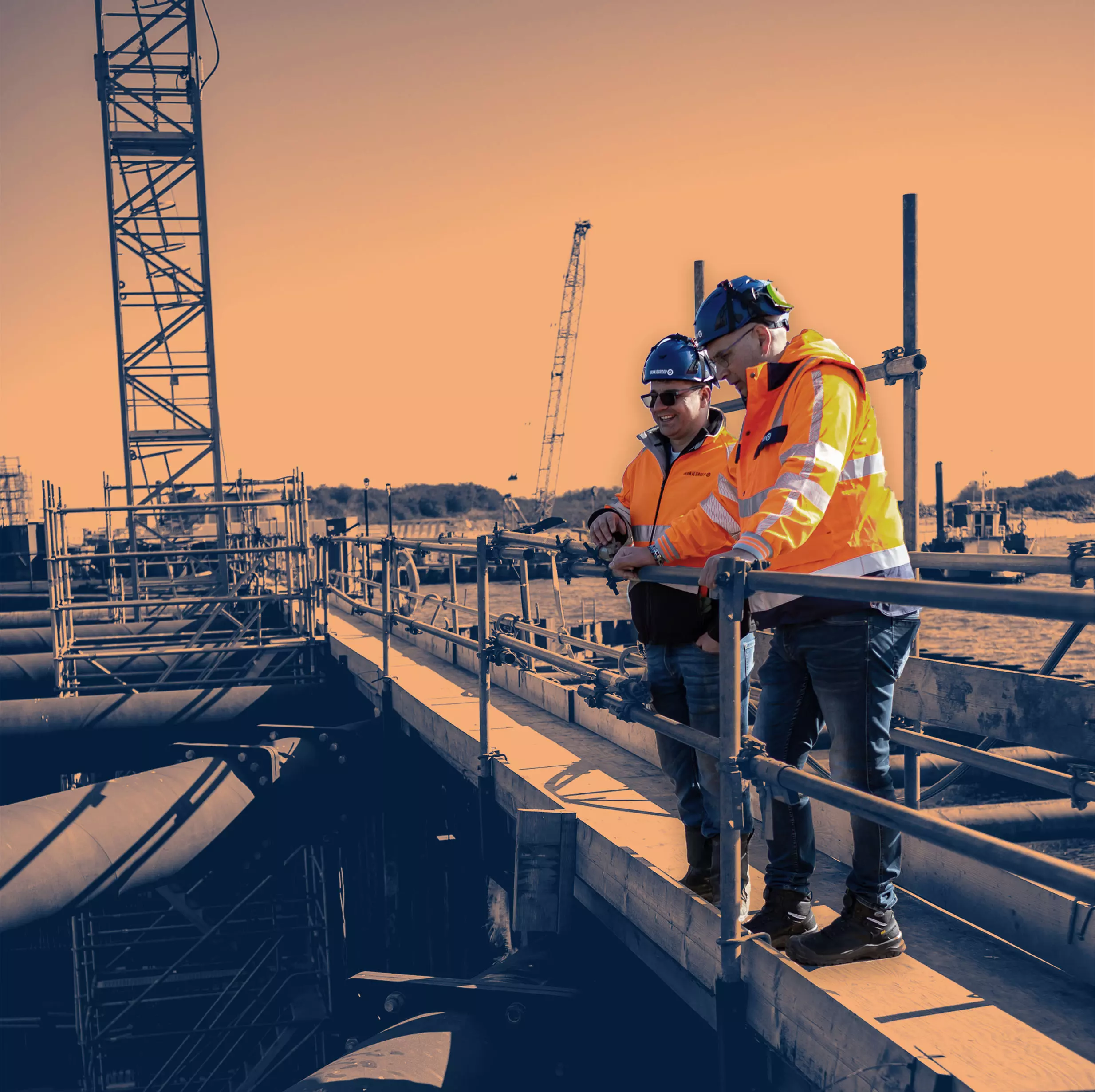 Two construction supervisors in safety gear discussing project progress on-site, standing on a platform above large industrial pipes.